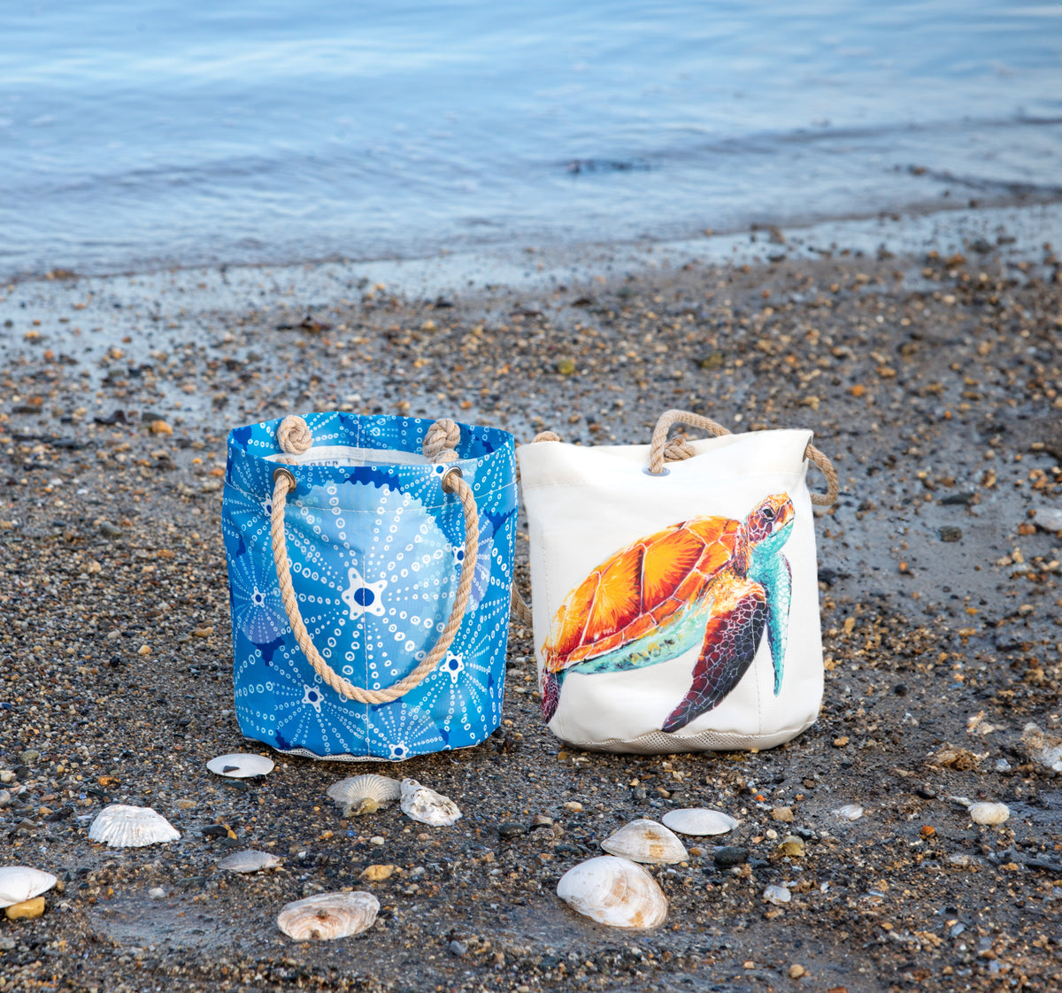 urchins bucket bag and watercolor sea turtle bucket bag perched on a pebbly beach