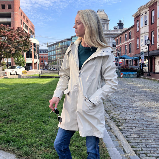 A woman wears a cream-colored, fleece-lined Harborside Waterproof Fleece-Lined Raincoat - Moonstruck while standing on a cobblestone street, prepared for coastal adventures.