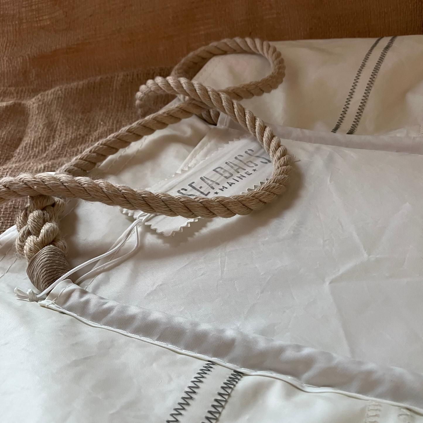 A close-up of the Sea Bags Wharf Tote, made from white sail cloth with rope handles and visible stitching, shown on a beige textured surface.