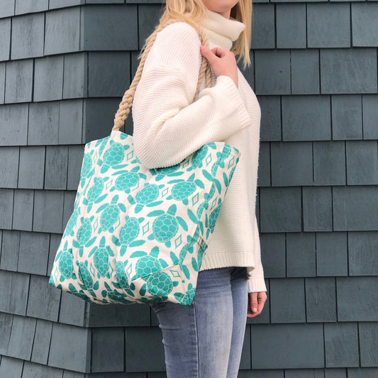 A person in a white sweater and jeans carries an Aquamarine Sea Turtle Medium Tote, a Sea Bags design made from recycled sail cloth with a teal turtle pattern and rope handles.
