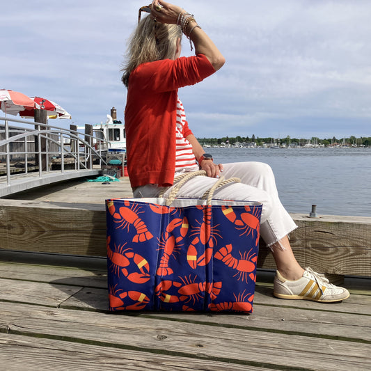 A woman sits on a wooden dock by the water wearing a red cardigan and white pants, with a navy blue lobster-print tote bag placed in front of her.