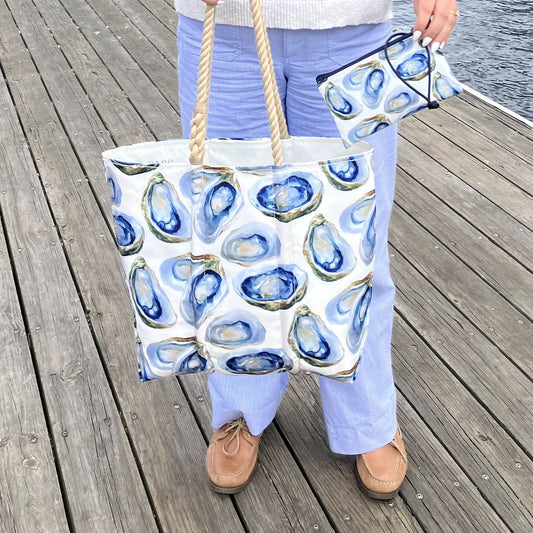 A person standing on a wooden dock holds a large white tote bag with blue oyster shell print and rope handles, while also holding a matching small zip pouch.