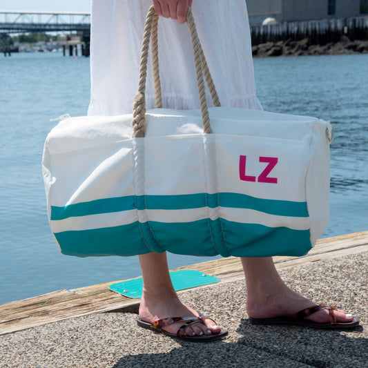 Wearing sandals and a white dress, a person stands on a dock by the water holding the Sea Bags Custom Monogram Breton Stripe Duffel Bucket Bundle, crafted from recycled sailcloth and featuring the initials “LZ” printed on it.