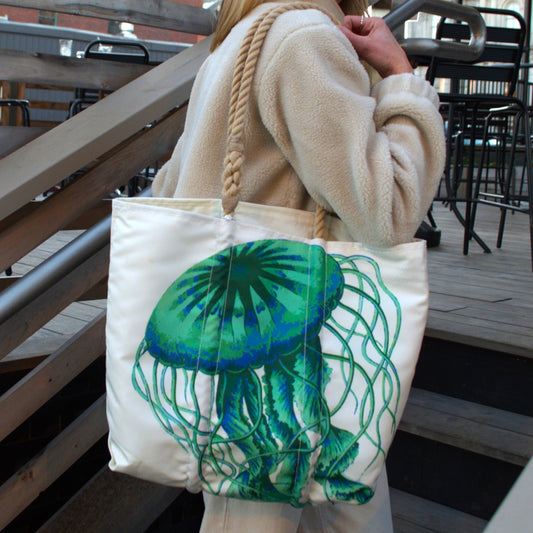 A person in a light-colored fleece jacket carries a Jellyfish Medium Tote by Sea Bags, featuring a large green and blue jellyfish graphic and rope handles.