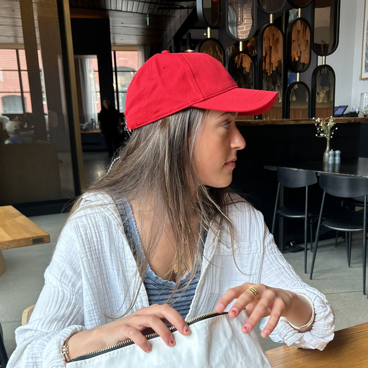 A woman wearing a bright red baseball cap, a white textured cardigan over a blue striped top, sitting in a modern cafe setting.
