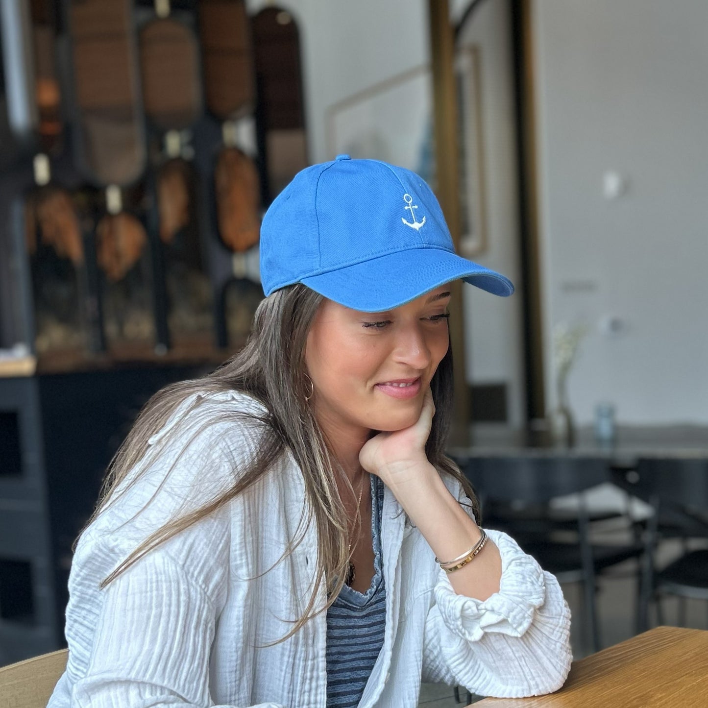 A woman wearing a bright blue baseball cap with a white embroidered anchor logo, resting her head on her hand at a wooden table in a canopy hotel setting.