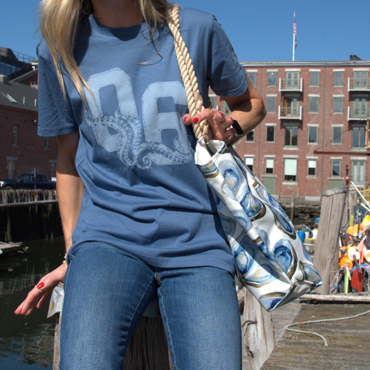 A woman wearing a blue graphic t-shirt and jeans stands on a wooden dock carrying a white tote bag with a blue and white oyster pattern and a thick rope handle, with a harbor and brick buildings in the background.