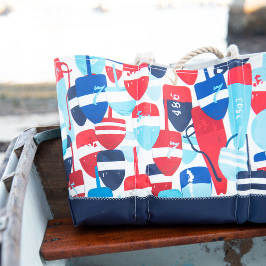 An Americana Buoys Large Tote by Sea Bags with a red, white, and blue buoy pattern and rope handles, sitting on a wooden bench inside a small boat.