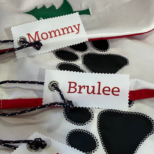 Three white dog bandanas with red trim and personalized names 'Mommy', 'Brulee', and 'Mez' on fabric tags, laid out on a tan surface.
