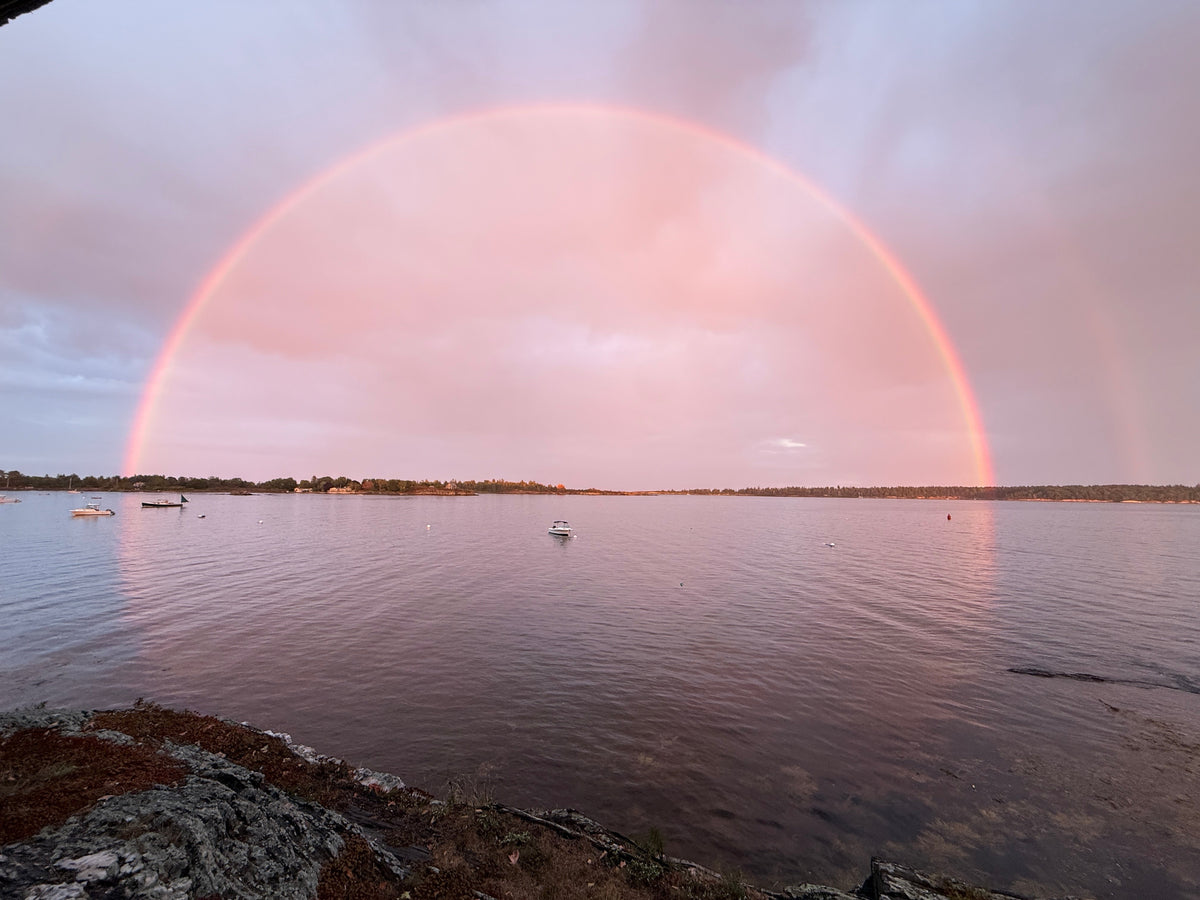 A wide rainbow arcs across a calm body of water at sunset, with a few small boats floating and distant tree-covered land under a partly cloudy sky.