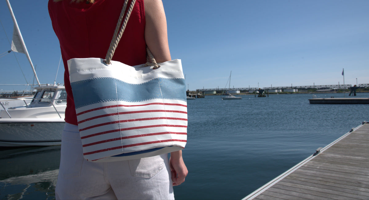 A woman viewed from behind carrying a large striped tote bag on a sunny day by the water, with boats and a dock visible in the background.