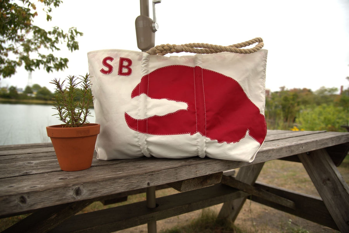 A person with long hair carries a large white tote bag featuring a blue lobster illustration. They stand in front of a wooden shingle wall with flowers in a white window box above.