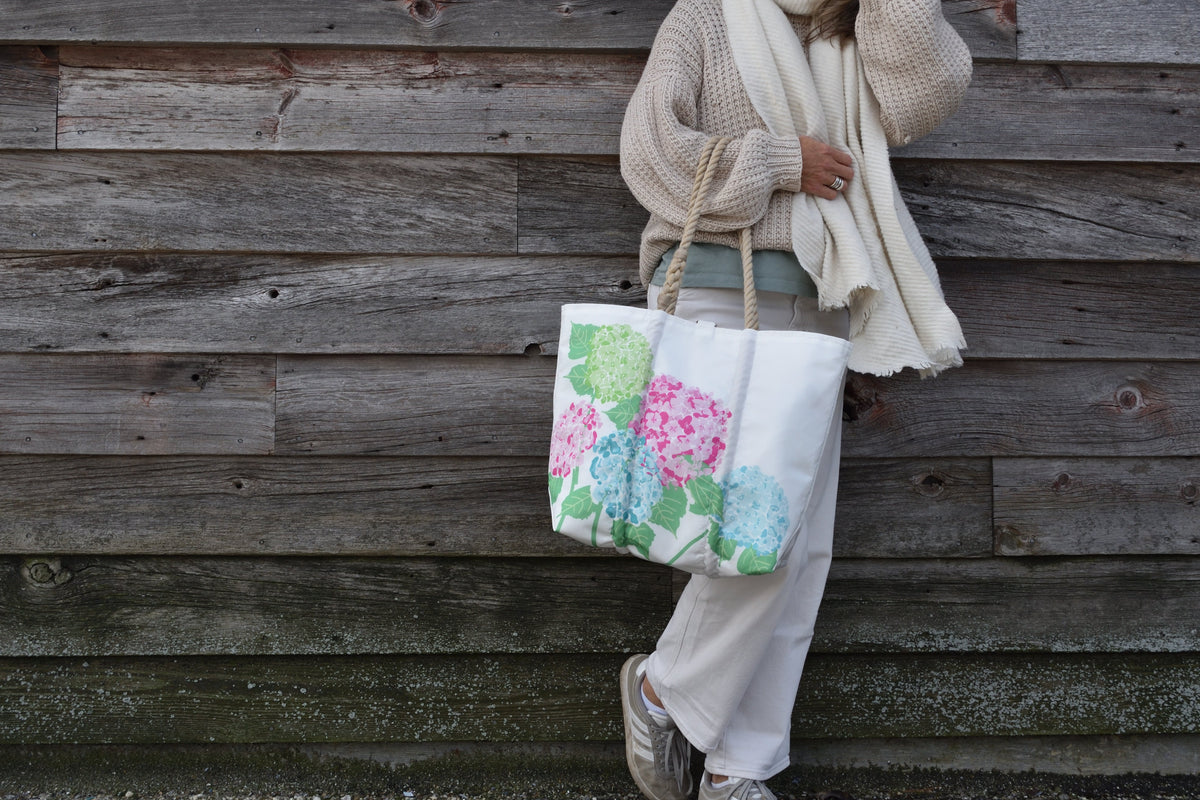 A person in a beige sweater and white pants carrying a white tote bag featuring a print of pink and blue hydrangea flowers.