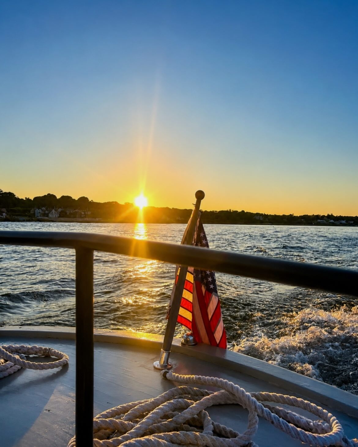 View from the deck of a boat with an American flag as the sun sets over the water.