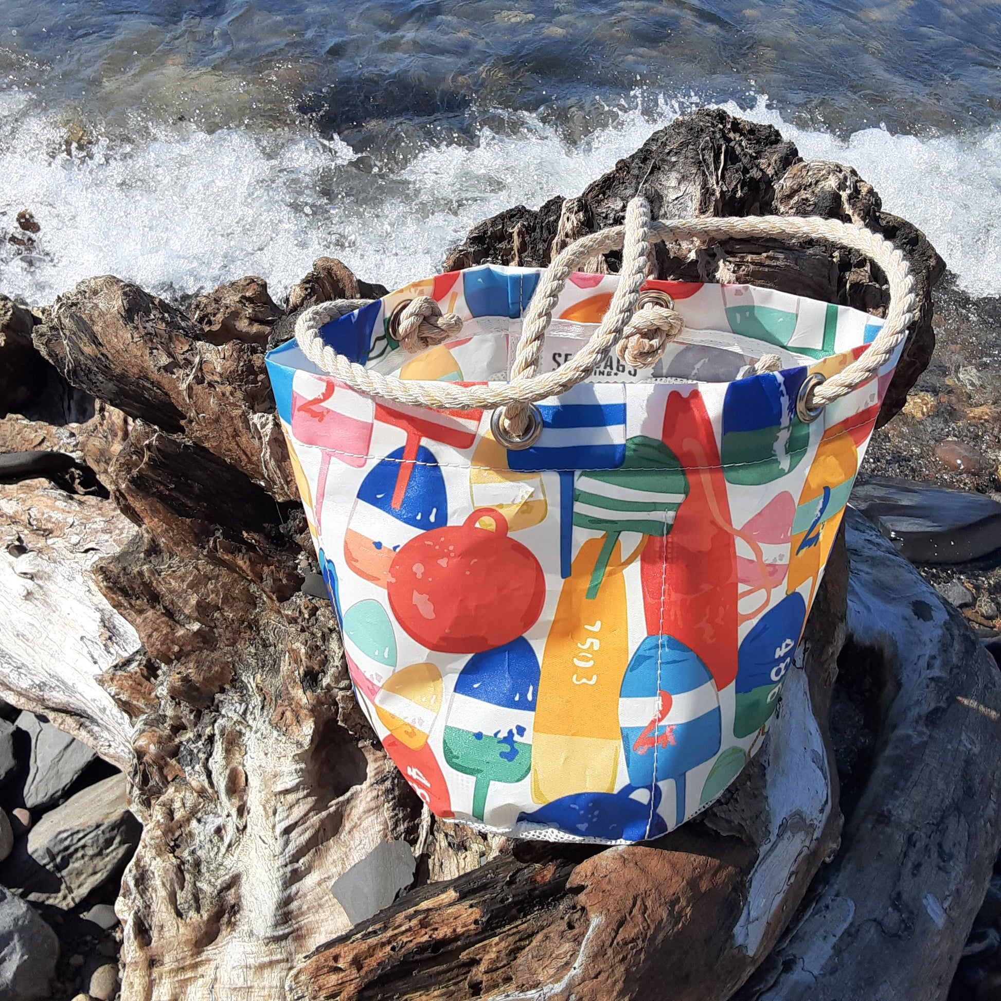 A tote bag with colorful nautical buoy illustrations and thick rope handles, resting on weathered driftwood with ocean waves crashing in the background.