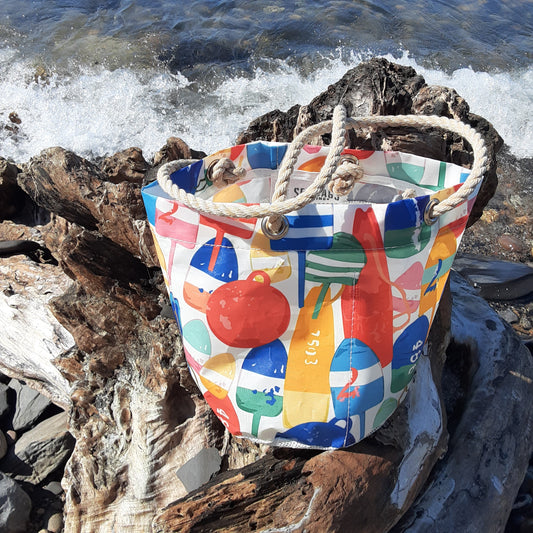 A tote bag with colorful nautical buoy illustrations and thick rope handles, resting on weathered driftwood with ocean waves crashing in the background.