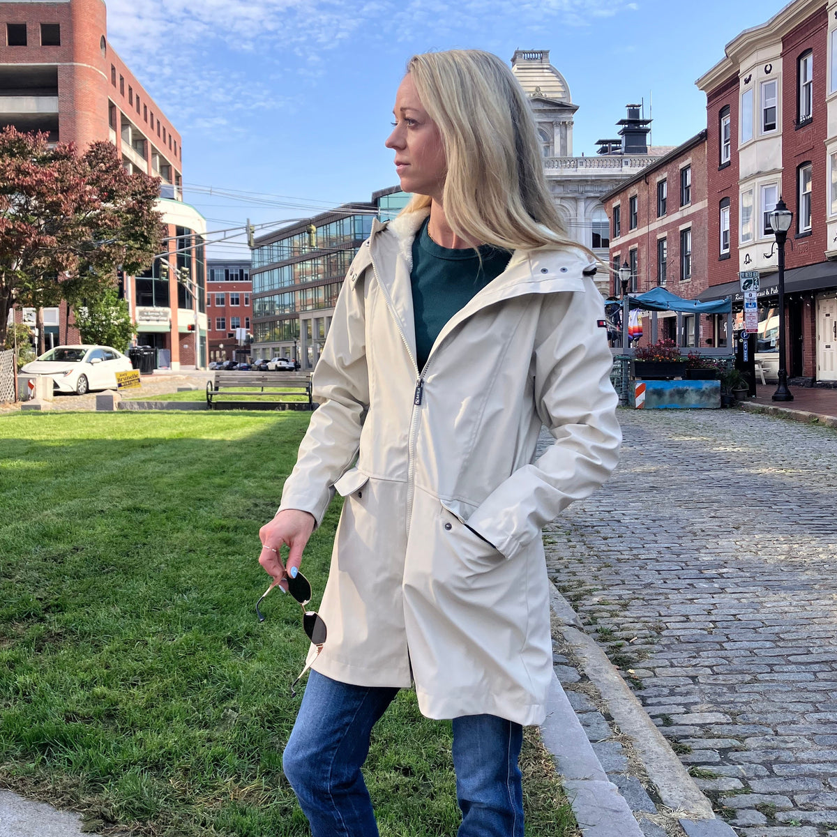 A woman wears a cream-colored, fleece-lined Harborside Waterproof Fleece-Lined Raincoat - Moonstruck while standing on a cobblestone street, prepared for coastal adventures.
