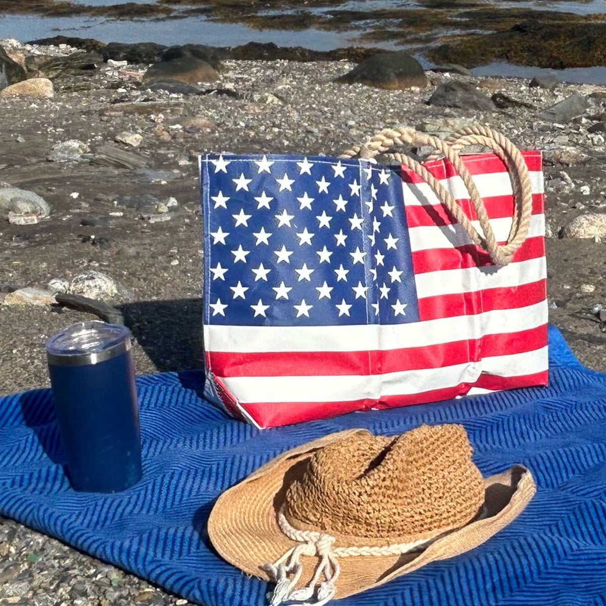 A Sea Bags Vintage Limited Edition American Flag Large Tote, a straw hat, and a metal tumbler rest on a blue mat on a rocky beach.