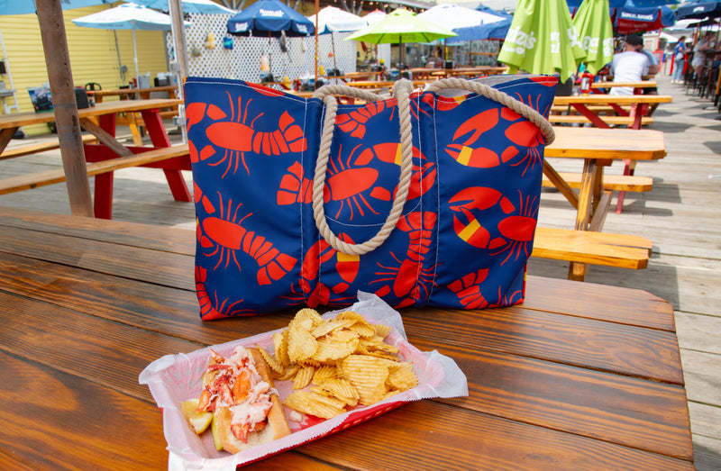 Outdoor dining scene on a wooden deck with two blue lobster-print tote bags, a matching bag holding iced beverage bottles, a yellow Butter bag, and a sandwich with chips and a dipping sauce on a wooden table.