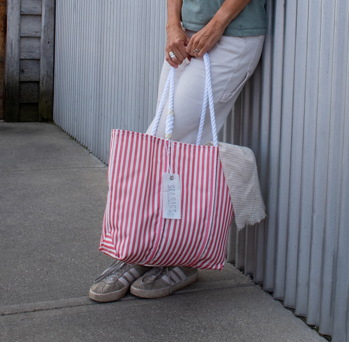 Person in white pants and sneakers stands by a corrugated wall, holding a Red Seersucker Stripe Medium Tote by Sea Bags—a nautical bag with rope handles and a tag attached.
