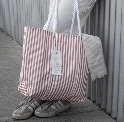 Person in white pants and sneakers stands by a corrugated wall, holding a Red Seersucker Stripe Medium Tote by Sea Bags—a nautical bag with rope handles and a tag attached.