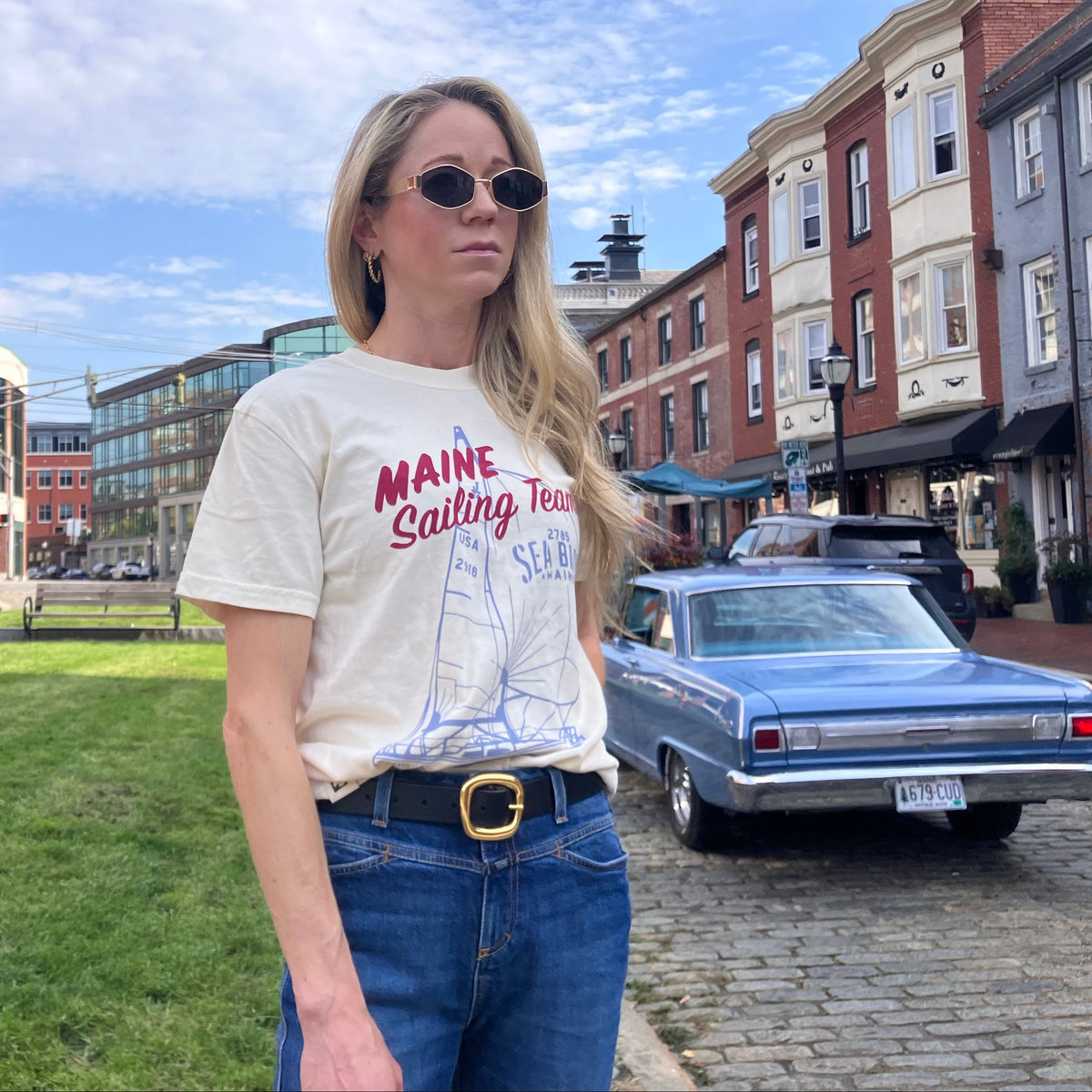 A woman wears a cream, ringspun cotton Maine Sailing Team T-Shirt featuring a blue and red nautical sailboat graphic in an outdoor urban setting.
