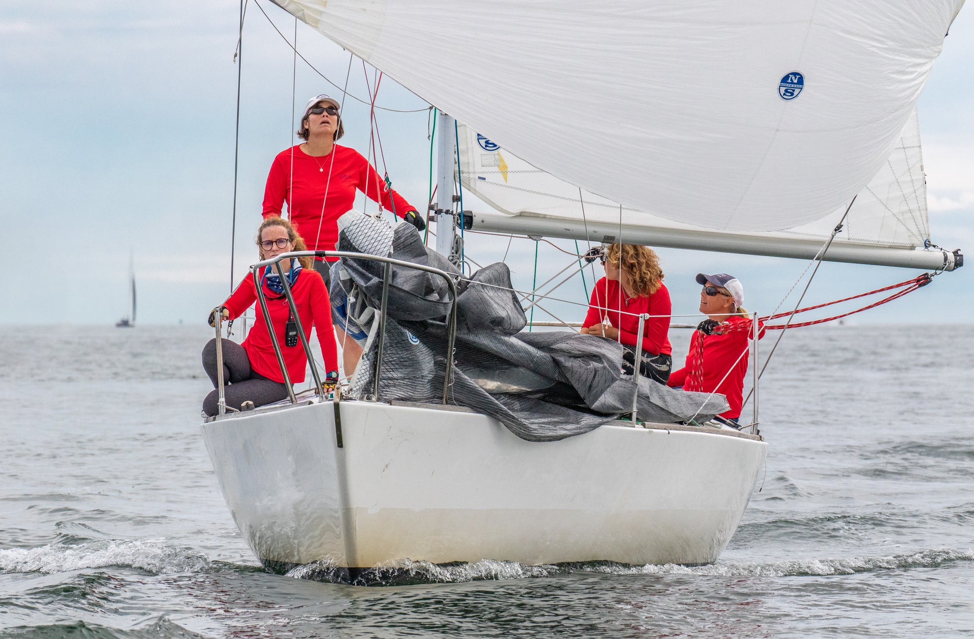 Four people wearing red shirts from the Sea Bags sail team navigate a small white sailboat on open water, sails up, with another boat visible in the distance.