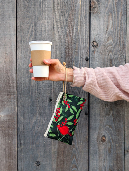 A person in a pink sweater holds a reusable coffee cup in one hand and the Sea Bags Winterberry Cardinals Wristlet—crafted from recycled sailcloth—in the other, against a wooden background. by Sea Bags