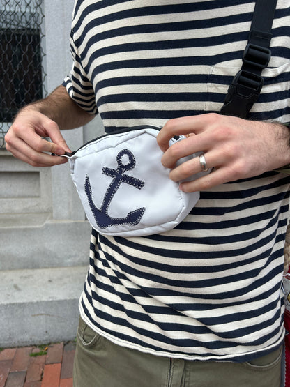 A person in a striped shirt opens the Sea Bags Navy Anchor Belt Bag, made from recycled sail cloth and featuring a navy blue anchor design, while standing in front of a stone building. by Sea Bags