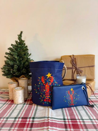 The Sea Bags Lobster Lights Bucket Bag, crafted from recycled sail cloth and featuring a delightful lobster motif, is displayed alongside its matching pouch on a plaid tablecloth. In the background, a mini Christmas tree, wrapped present, and birch candles evoke a cozy New England maritime holiday ambiance. by Sea Bags