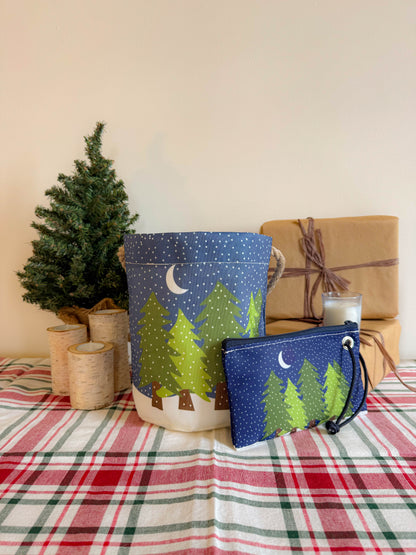 A small tree, birch candles, a wrapped gift, and two bags featuring a night forest design—including the Sea Bags Midnight Snow Bucket Bag made from recycled sail cloth—rest on a plaid tablecloth. by Sea Bags