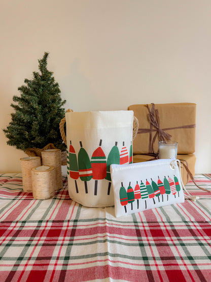 A holiday-themed table featuring a plaid cloth, small pine tree, three candles, a wrapped gift, and festive bags and pouches adorned with green and red trees creates a cozy coastal vibe—especially with the Sea Bags Holiday Buoys Bucket Bag, crafted from recycled sail cloth, adding nautical charm to the scene. by Sea Bags