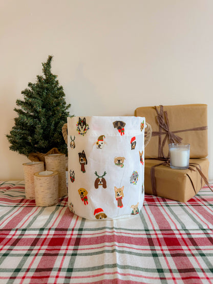 The Very Merry Pups Bucket Bag by Sea Bags, adorned with delightful dog illustrations, rests on a plaid tablecloth beside a small artificial tree, candles including one in a glass, and two wrapped gifts—offering an inviting and festive touch of holiday décor for any dog lover. by Sea Bags