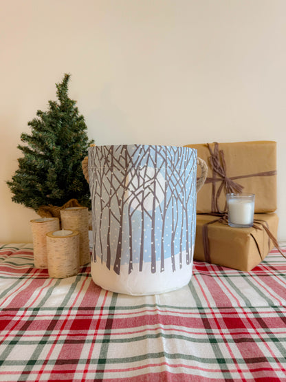 The Winter Solstice Bucket Bag from Sea Bags, crafted from recycled sail cloth and featuring a winter tree design, is displayed on a plaid tablecloth next to a small tree, candles, and two wrapped gifts. by Sea Bags
