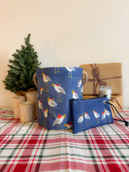 The Caroling Gulls Bucket Bag by Sea Bags, featuring bird designs evocative of coastal Christmas decor, sits on a plaid tablecloth beside two candles, a small tree, and a wrapped gift. by Sea Bags