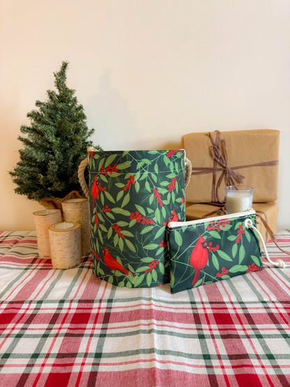 A small artificial tree, three birch-style candle holders, and wrapped gifts are displayed on a plaid tablecloth with a matching green floral pouch in the foreground, alongside the Sea Bags Winterberry Cardinals Bucket Bag made from sustainable recycled sail cloth for charming holiday decor. by Sea Bags