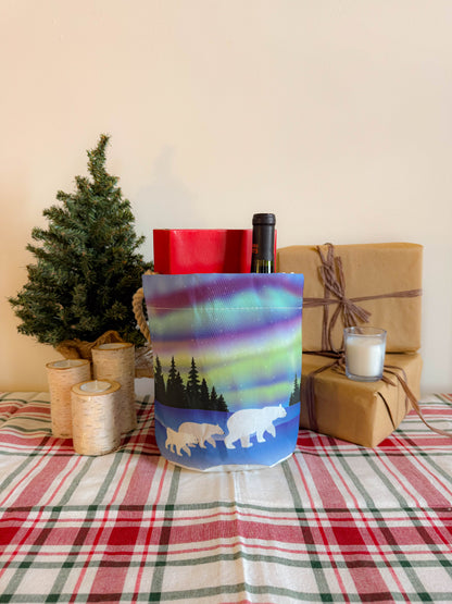 Celebrate the holidays with the Sea Bags Northern Lights Bucket Bag, featuring a charming polar bear family design on a plaid tablecloth beside candles, a mini Christmas tree, and wrapped gifts—crafted from recycled sail cloth for an eco-friendly festive touch. by Sea Bags