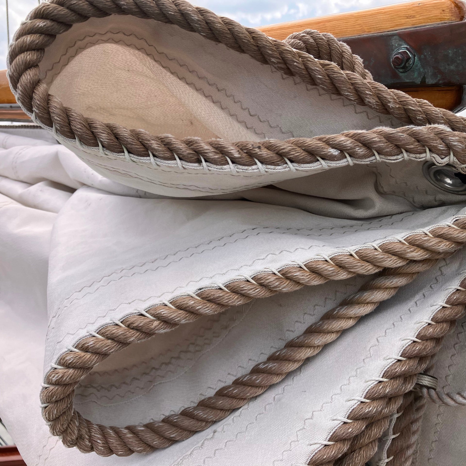 Close-up of a folded sail with thick, twisted rope edging and visible stitching, reminiscent of the craftsmanship found in Sea Bags; part of a wooden boat is seen in the background.