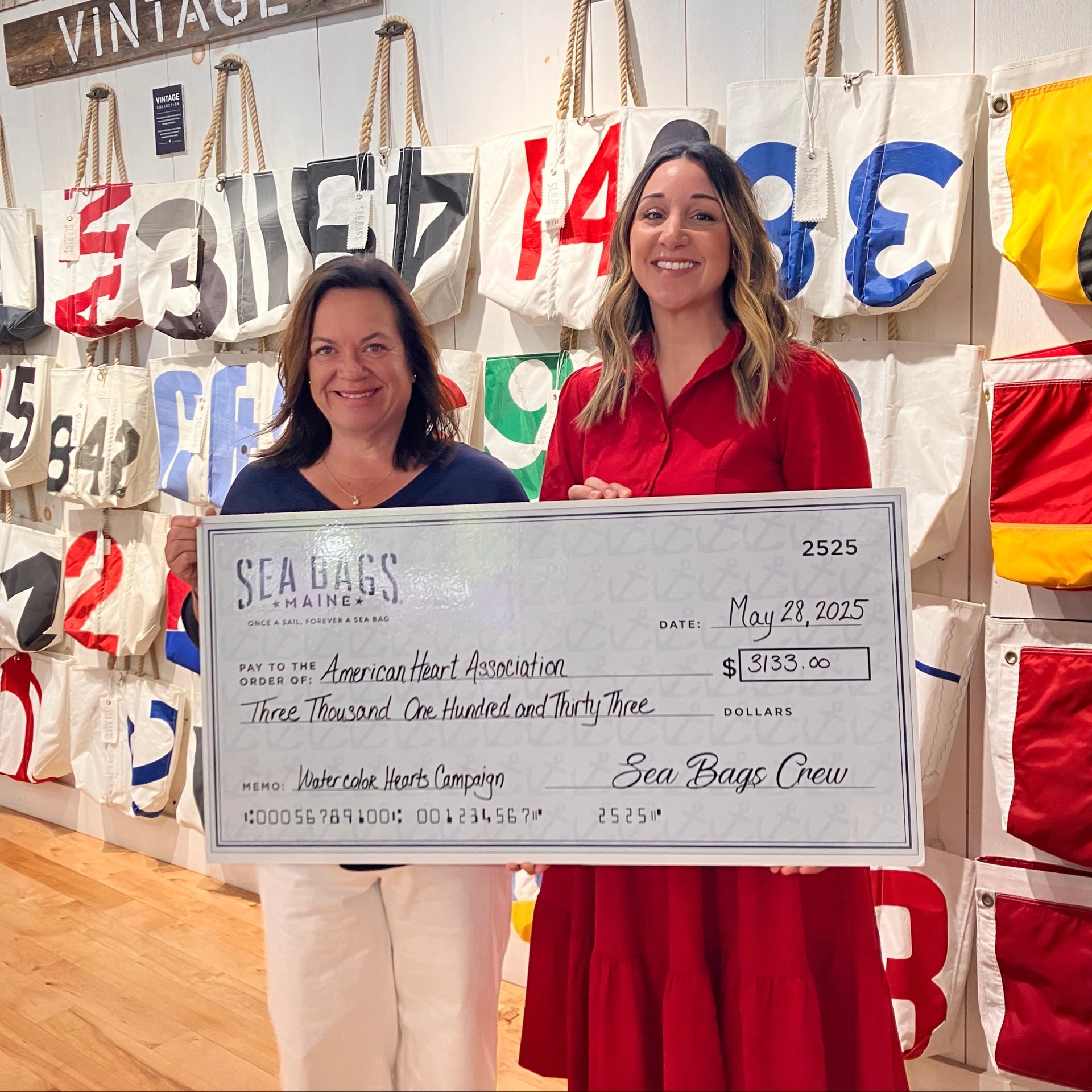 Sea Bags CEO, Beth Greenlaw, stands indoors holding a donation check to the American Heart Association, surrounded by colorful tote bags with nautical designs on the wall.