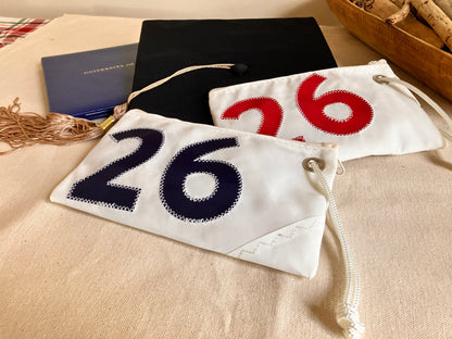 Two small Navy 2026 Graduation Wristlets from Sea Bags, crafted from recycled sail cloth and featuring the number 26 sewn on them in blue and red, are placed next to a graduation cap and tassel on a beige fabric surface—making for a thoughtful 2026 graduate gift or earth-friendly organizer. by Sea Bags