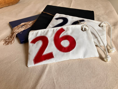 The Sea Bags Red 2026 Graduation Wristlet, crafted from recycled sail cloth and adorned with a bold red 2026, rests on beige fabric beside a graduation cap with a gold tassel and a dark blue folder—making it a distinctive and meaningful gift for any special occasion. by Sea Bags
