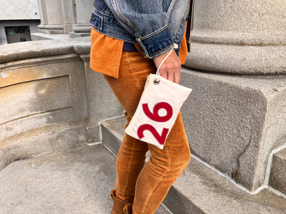 A person in orange corduroy pants and a denim jacket stands by a stone wall, holding the Sea Bags Red 2026 Graduation Wristlet—crafted from recycled sail cloth and featuring a bold red 26 on white. by Sea Bags