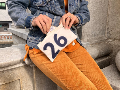 Person in orange corduroy pants and a denim jacket puts a folded bill into a Navy 2026 Graduation Wristlet by Sea Bags—an earth-friendly organizer crafted from recycled sail cloth—while sitting on a stone ledge. by Sea Bags