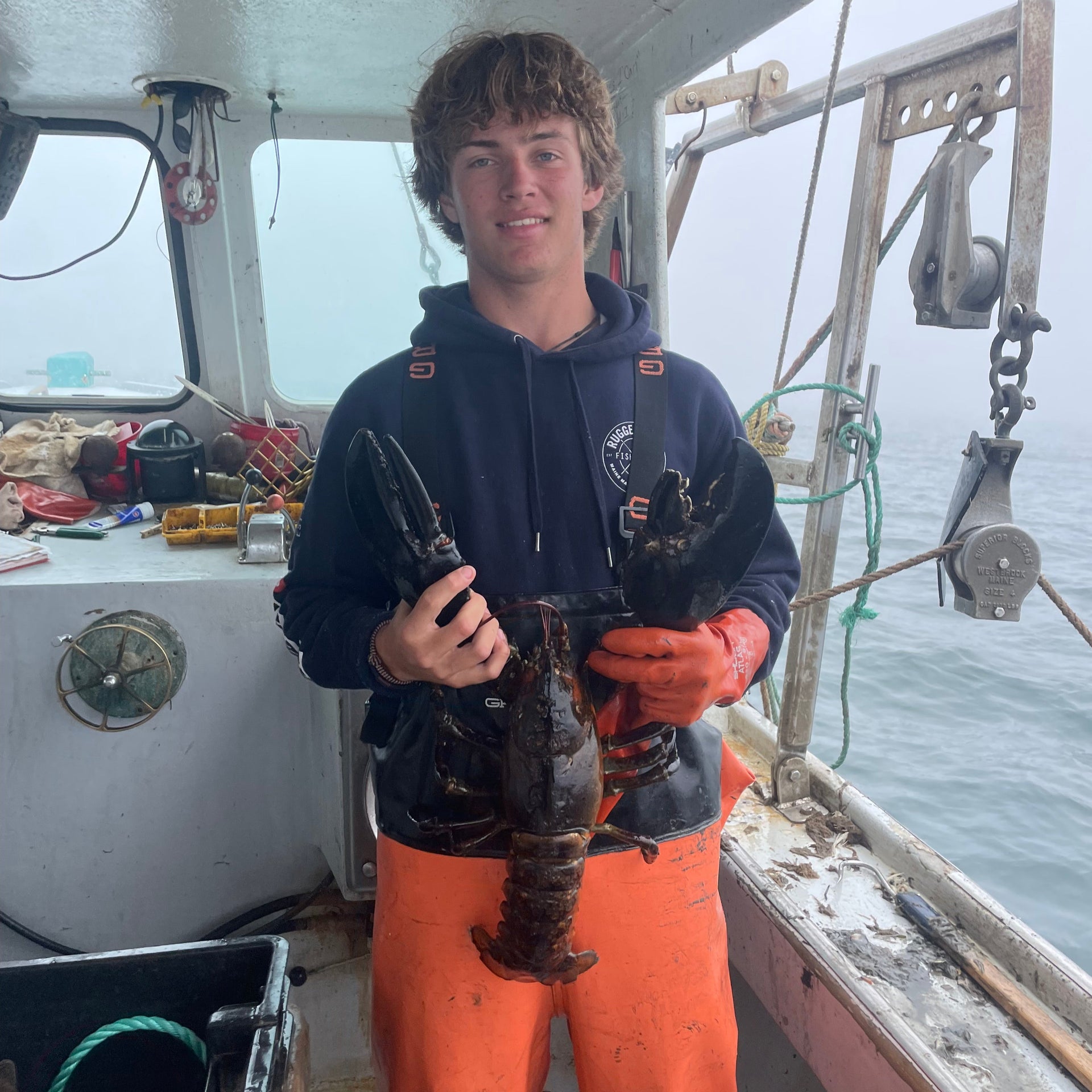 A young fisherman wearing orange overalls and gloves holds a large lobster on a fishing boat, with equipment and the ocean visible in the background.