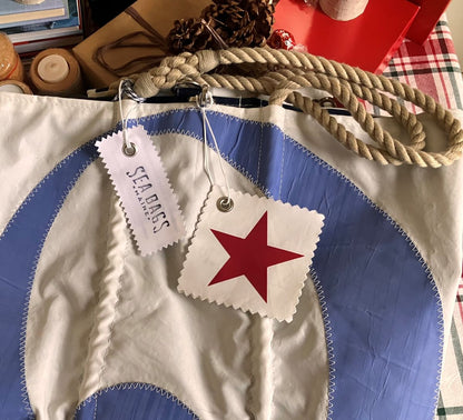 Elevate your holiday gifting with the Sea Bags Star Sail Cloth Gift Tag, featuring a unique red star and blue circle design on recycled sailcloth, accented by rope handles and two attached tags—one branded with Sea Bags and the other signifying its eco-friendly origins as a recycled sail. by Sea Bags