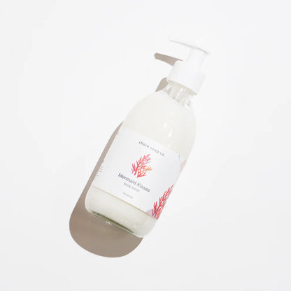 Clear glass pump bottle labeled “Mermaid Kisses Hand Soap” by Shore Soap Co., with a red coral graphic, sits beside Mermaid Kisses Eco-Friendly Body Lotion on a white background.
