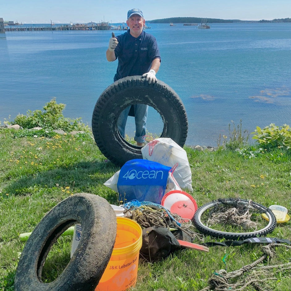 A Sea Bags reclaimer stands behind a large tire and gives a thumbs up near a waterfront, surrounded by collected trash including tires, ropes, buckets, and bags.