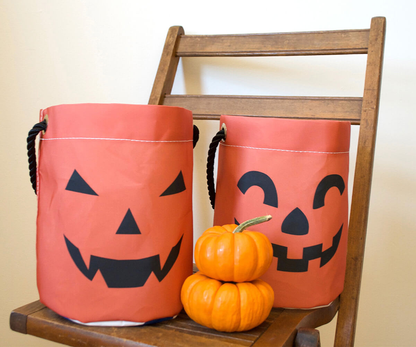 Two Sea Bags Jack O’Lantern Bucket Bags with cheerful jack-o’-lantern faces rest on a wooden chair beside a small stack of two mini pumpkins, all set for a festive night of trick-or-treating.
