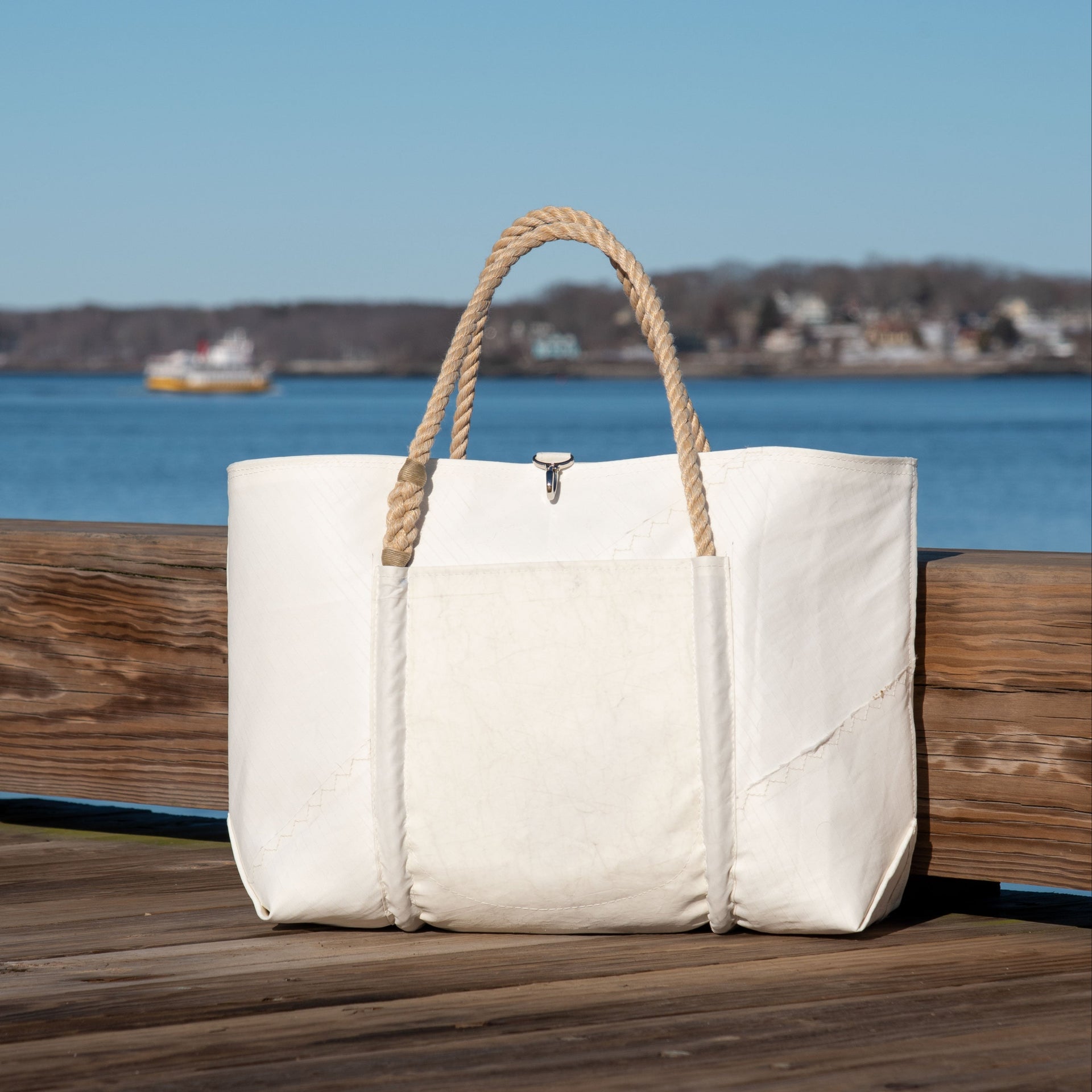 A large white Jeb tote bag with rope handles sits on a wooden dock, with water and a distant shoreline in the background, capturing the coastal style Sea Bags are known for.