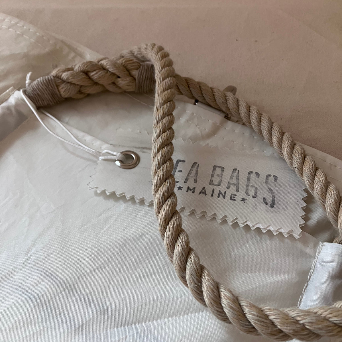 Close-up of a white fabric bag with a thick twisted beige rope handle and a sewn-on brand tag reading SEA BAGS MAINE.